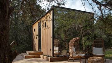 Uma cabana de madeira com porta de vidro, rodeada por uma floresta. Há cadeiras de vime e uma pequena mesa do lado de fora.