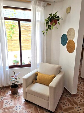Ein Zimmer mit einem beigen Sessel, einem Blumentopf, einer Wanduhr und einem Fenster mit Blick nach draußen.
