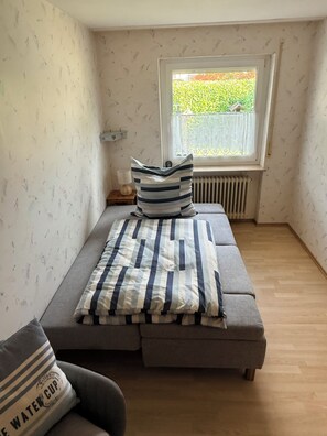 A small, cozy room with a grey sofa, a window with a view of greenery, and a radiator under the window. - Casa Ruf - Near Europa-Park (Hohberg)