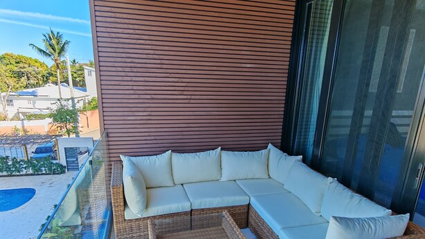 A modern outdoor seating area with a wicker sofa and cushions, a glass balustrade, and a view of a residential area. - Surf One (Sosúa)