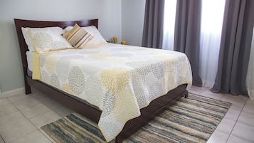 Una cama con colcha estampada, una mesita de noche con lámpara y una ventana con cortinas.