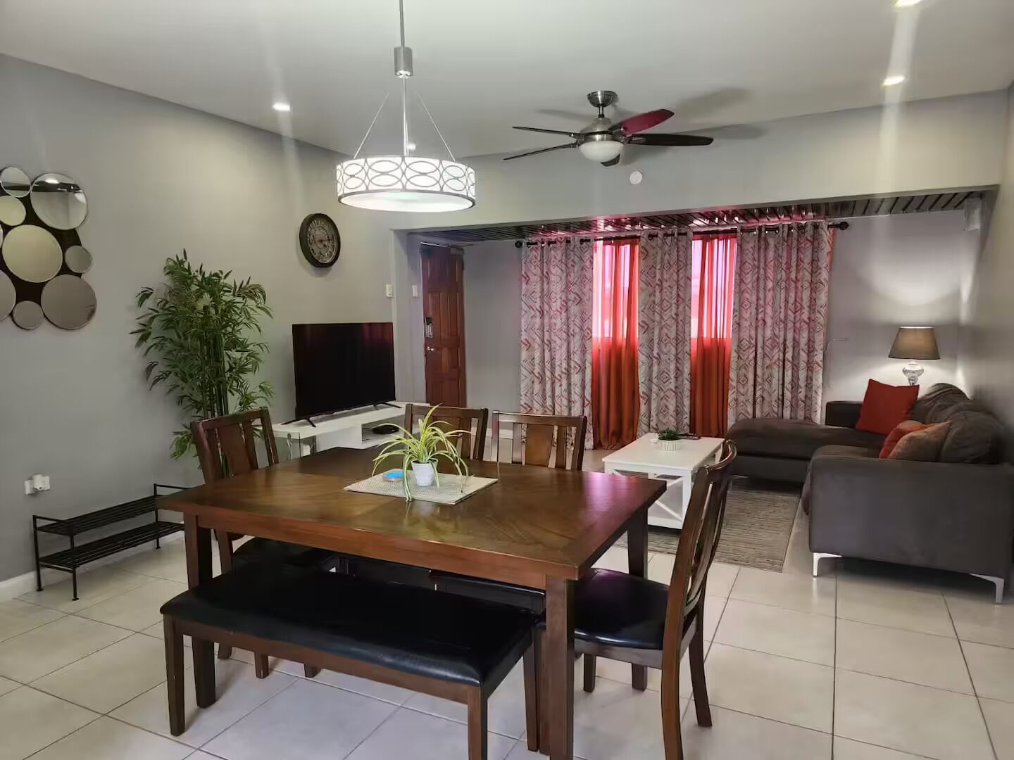 A modern living room with a dining area, a flat-screen TV, a ceiling fan, and a large window with curtains.
