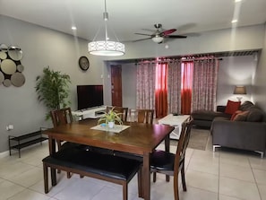 A modern living room with a dining area, a flat-screen TV, a ceiling fan, and a large window with curtains.
