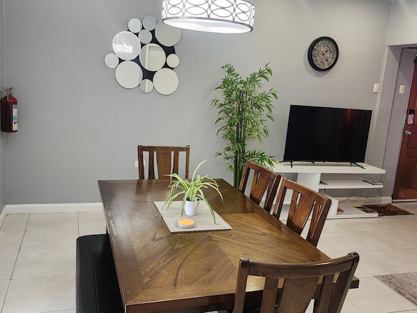A dining area with a wooden table, chairs, a potted plant, a wall clock, and a television on a stand.