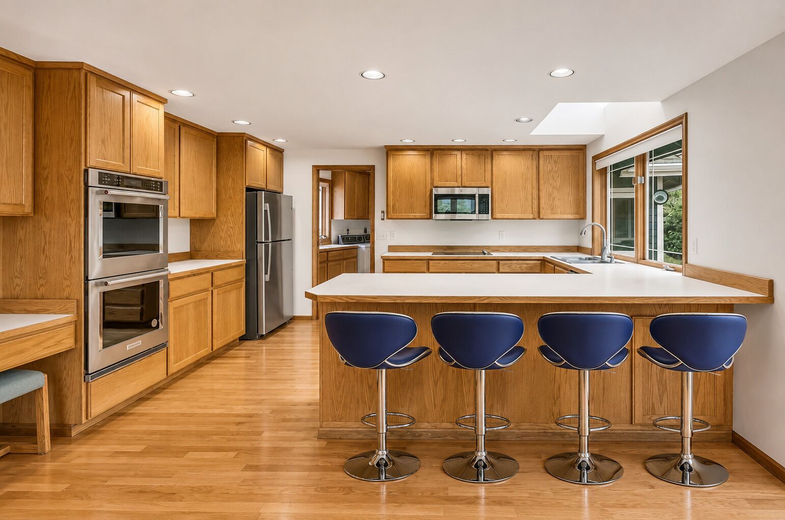 A modern kitchen with wooden cabinets, stainless steel appliances, a central island with bar stools, and large windows.