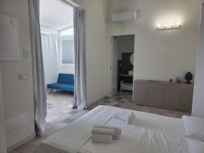 Ein Hotelzimmer mit einem Bett, einem blauen Sessel, einem Schminktisch mit Spiegel und einem durch eine offene Tür sichtbaren Badezimmer.