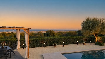 Zona de piscina con una pérgola, mobiliario exterior y vistas a un paisaje urbano al atardecer.