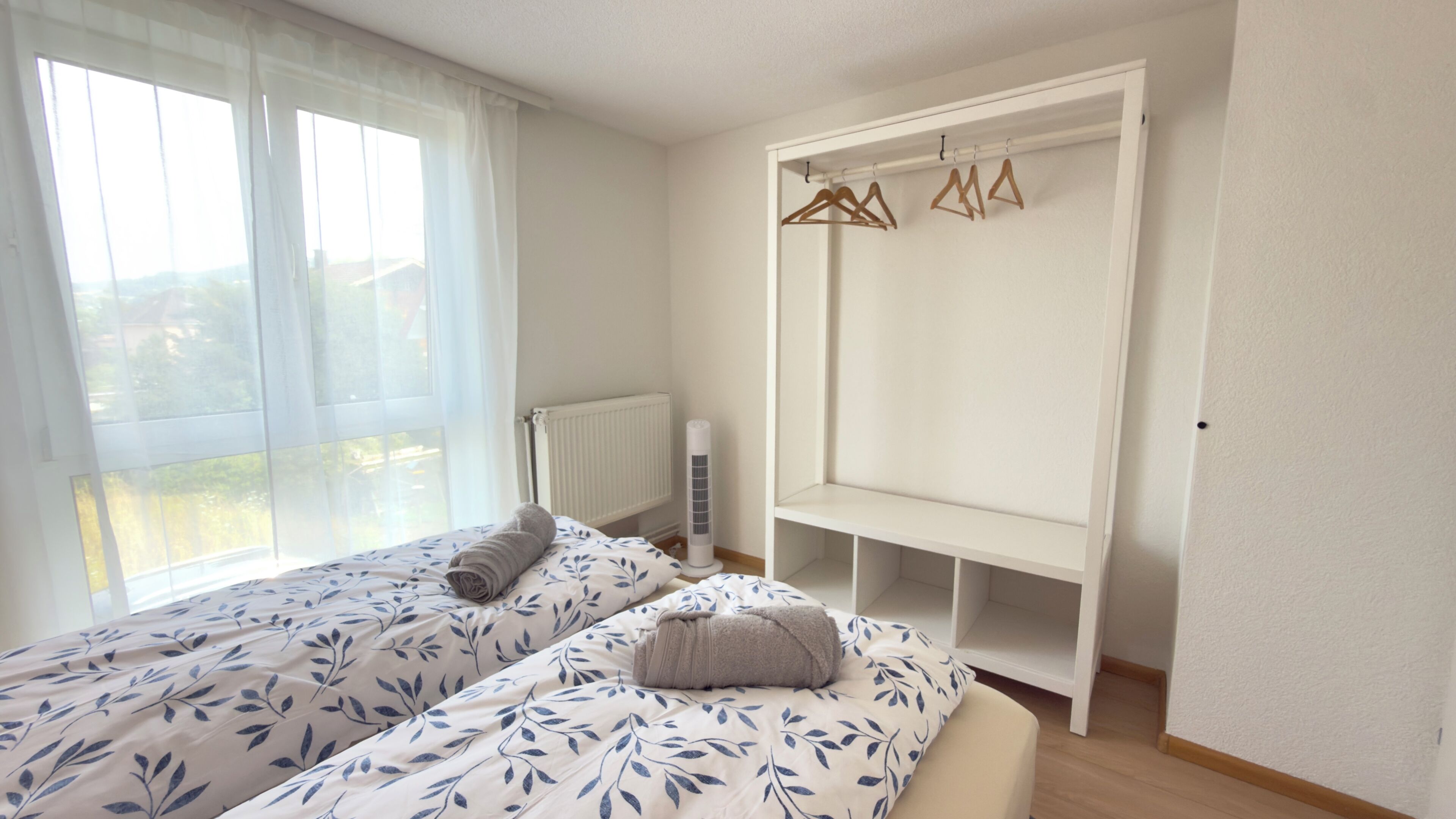 Ein Schlafzimmer mit zwei Betten, einem Heizkörper, einem weißen Kleiderschrank mit Kleiderhaken und einem Fenster mit durchscheinenden Vorhängen.