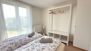 Un dormitorio con dos camas, un radiador, un armario blanco con percheros y una ventana con cortinas traslúcidas.