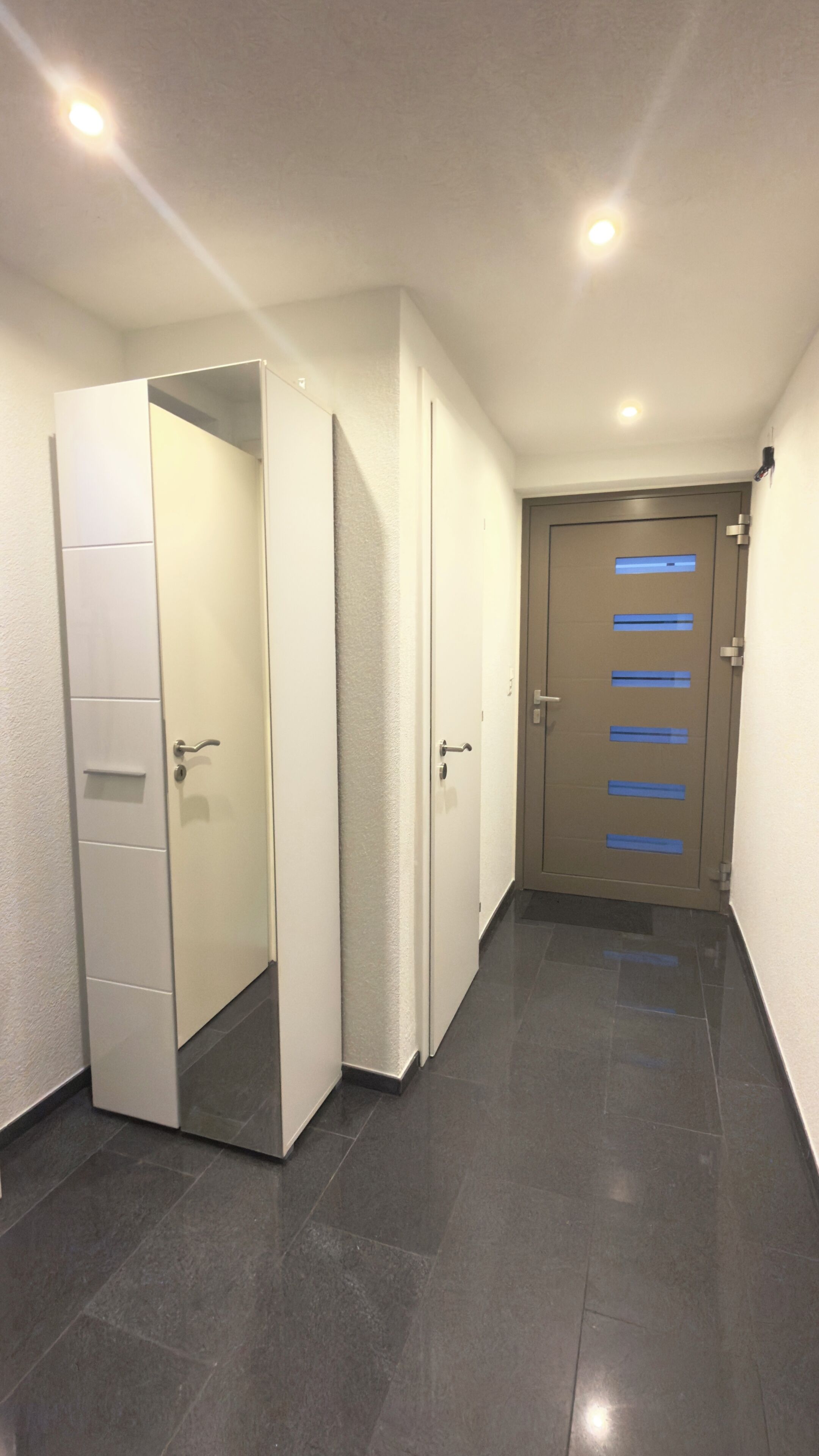 A hallway with a closed door at the end, a white wardrobe with a mirror, and a tiled floor.