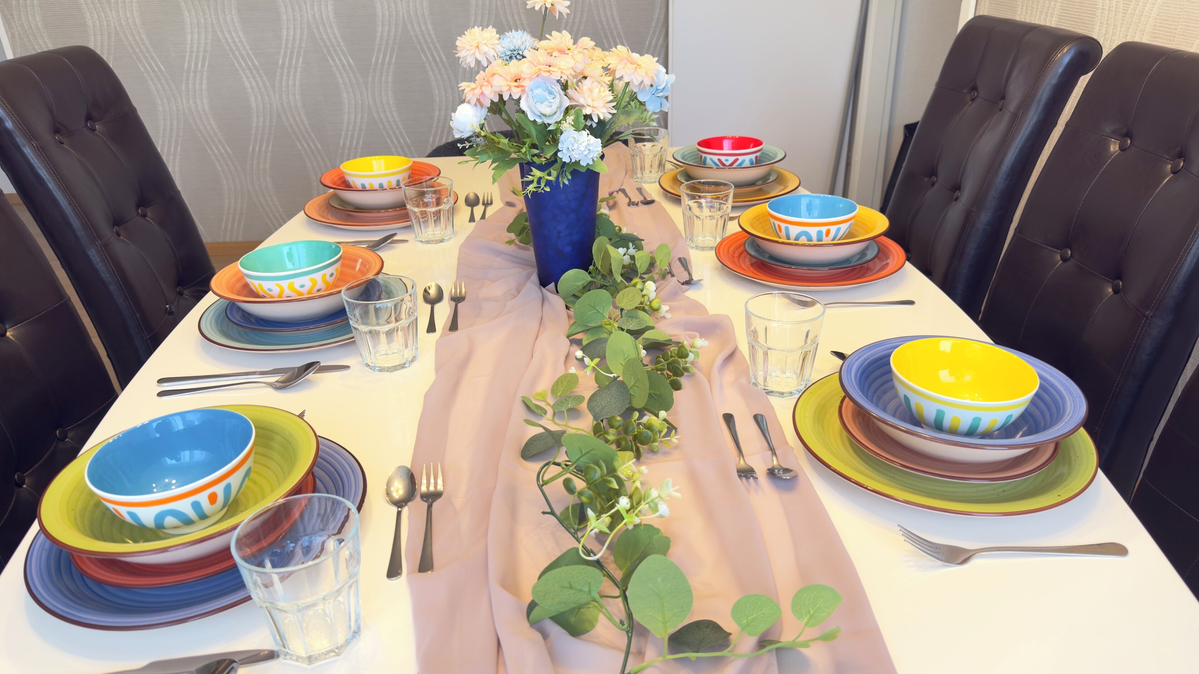 Une table à manger dressée avec des bols, des verres et des couverts aux couleurs vives, entourée de chaises en cuir noir.