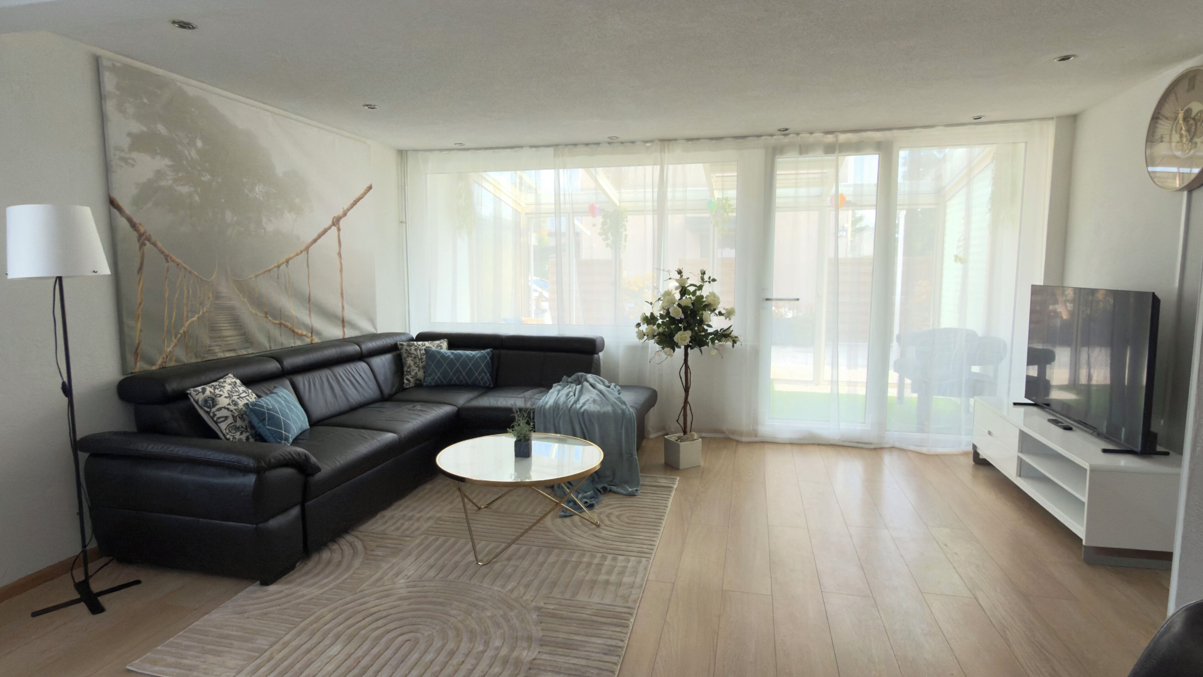 Un salon moderne avec un canapé d’angle, une table basse ronde, un téléviseur et une grande fenêtre avec des rideaux.