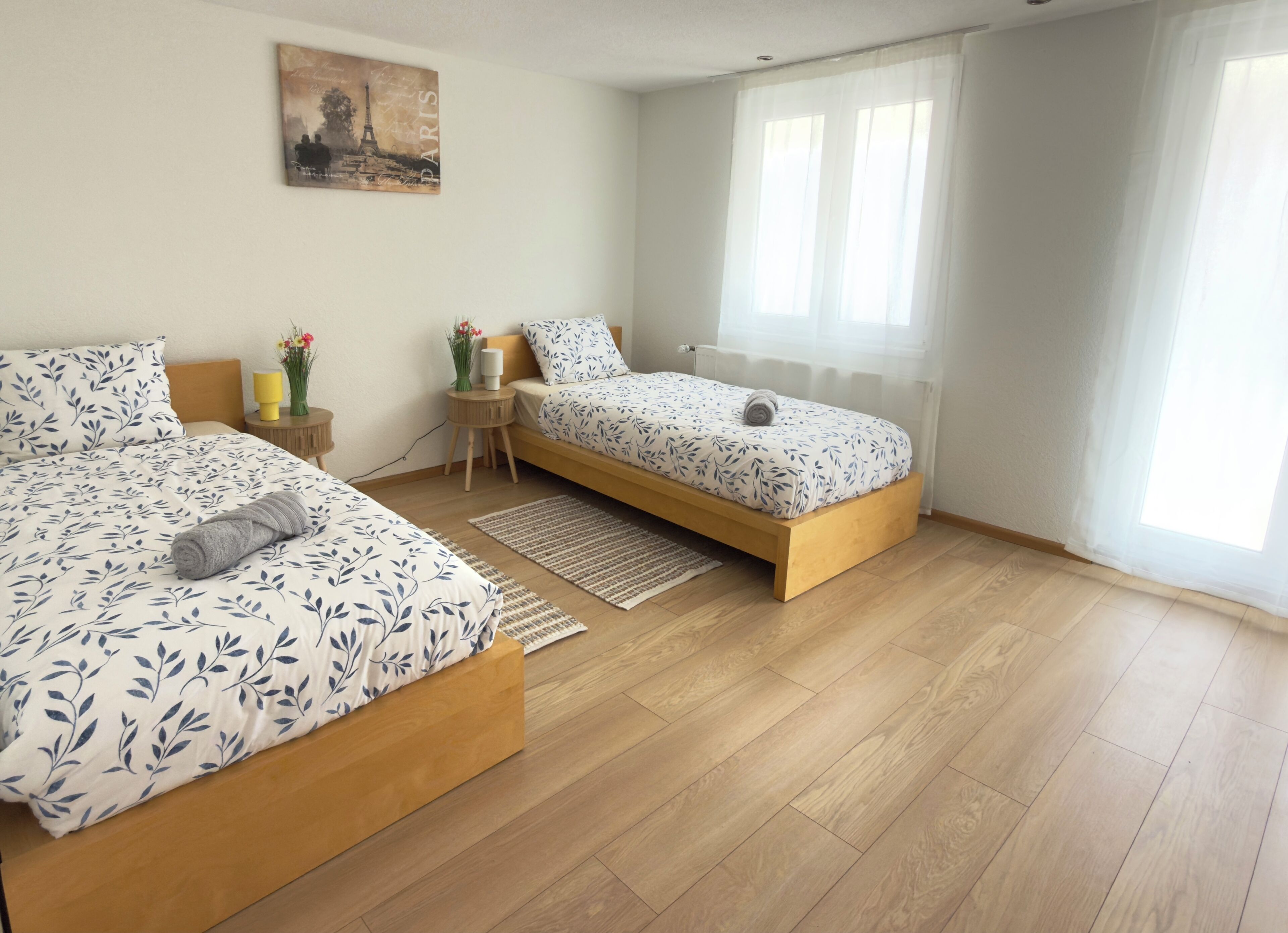 A bedroom with a large bed, a smaller bed, a nightstand, a lamp, and a window with sheer curtains.