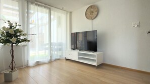 A modern living room with a flat-screen TV on a white stand, a potted plant, and a sliding glass door leading to an outdoor area.