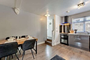 A modern kitchen with a dining area, featuring a built-in oven, a stove, and a window with blinds. - Host & Stay | The Coach House (Worthing)