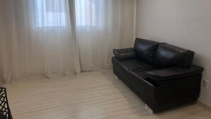 Una sala de estar con un sofá de cuero negro, cortinas claras y suelo de madera clara.