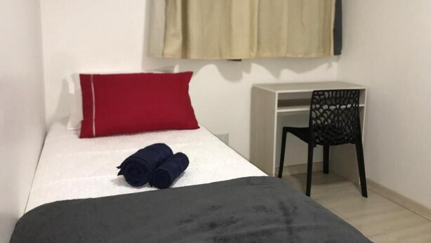 Una cama individual con un cojín rojo y toallas azules oscuras enrolladas, una silla negra y un escritorio blanco en una habitación con una cortina beige.