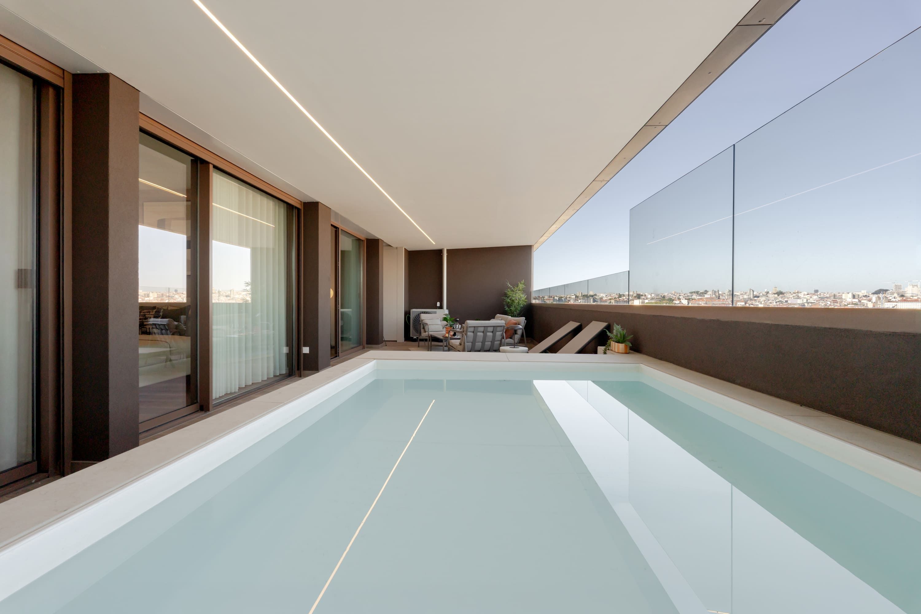 Area piscina interna moderna con vista sulla città, ampie porte scorrevoli in vetro e una zona relax con tavolo e sedie.