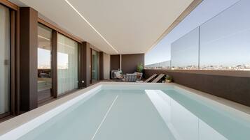 Area piscina interna moderna con vista sulla città, ampie porte scorrevoli in vetro e una zona relax con tavolo e sedie.