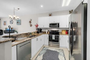 A modern kitchen with white cabinets, stainless steel appliances, a granite countertop, and a chandelier. - World Tour Fairway Condo • Pool • 10 Min to Beach (Myrtle Beach)