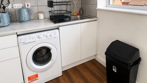Uma cozinha compacta com armários brancos, máquina de lavar, pia e um cesto de lixo preto.