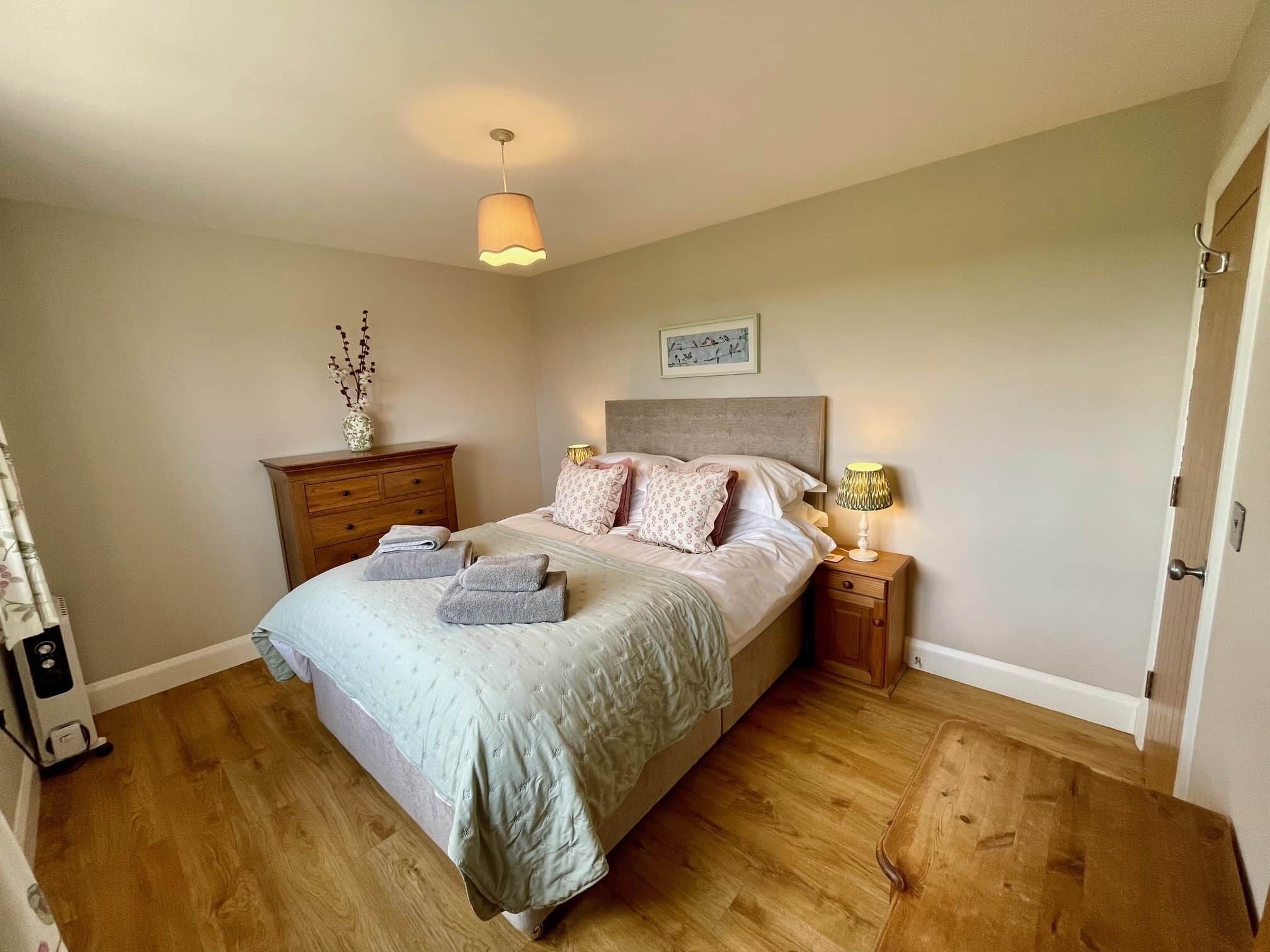 A bedroom with a bed, bedside tables, a nightstand, a lamp, a picture on the wall, and a vase with branches.