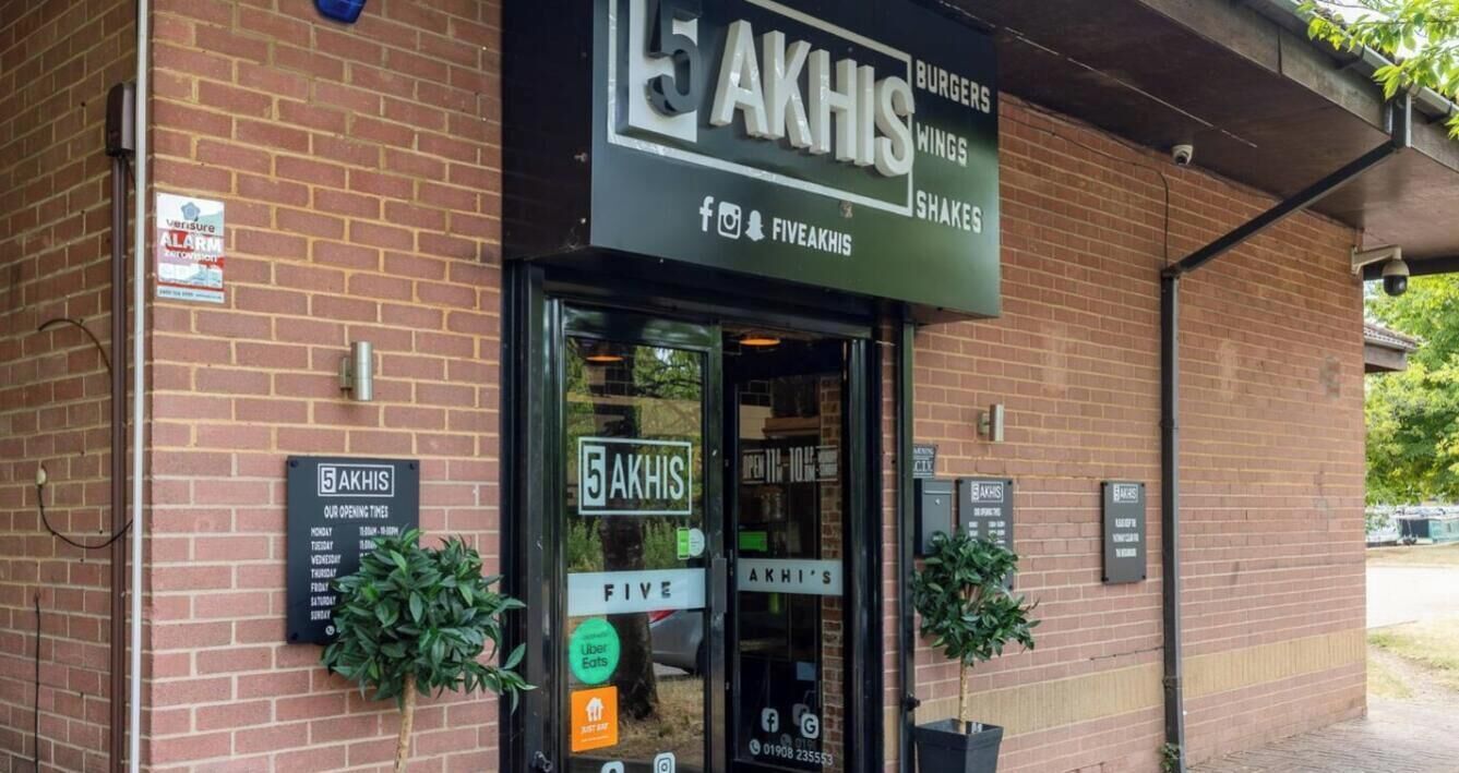 Un bâtiment en briques surmonté d’un auvent noir où l’on peut lire l’inscription « 5 A K H I S », ainsi que des mentions concernant les hamburgers, les ailes de poulet et les milk-shakes.