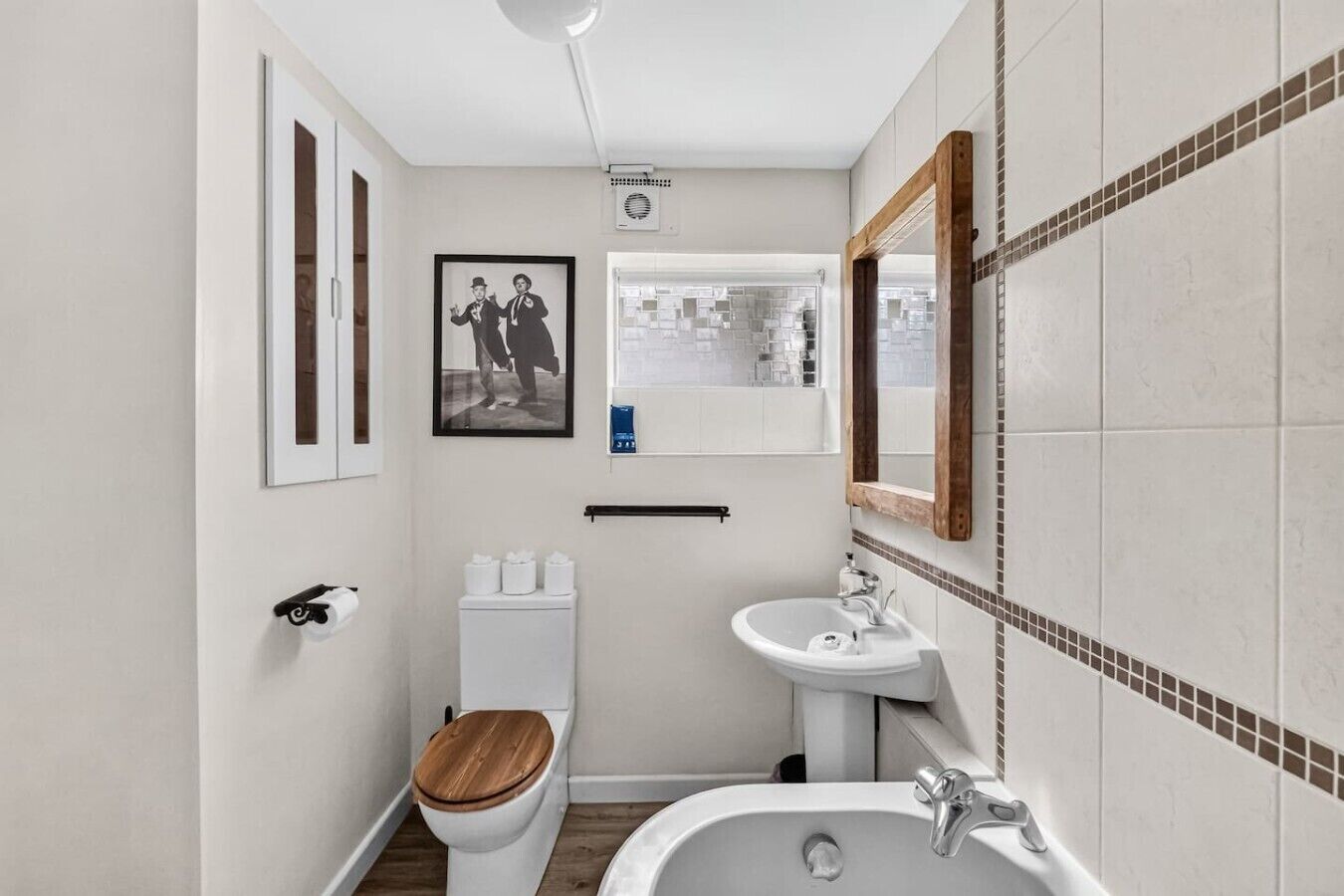 A compact bathroom with a toilet, sink, and bathtub. There is a framed picture on the wall and a window above the sink.