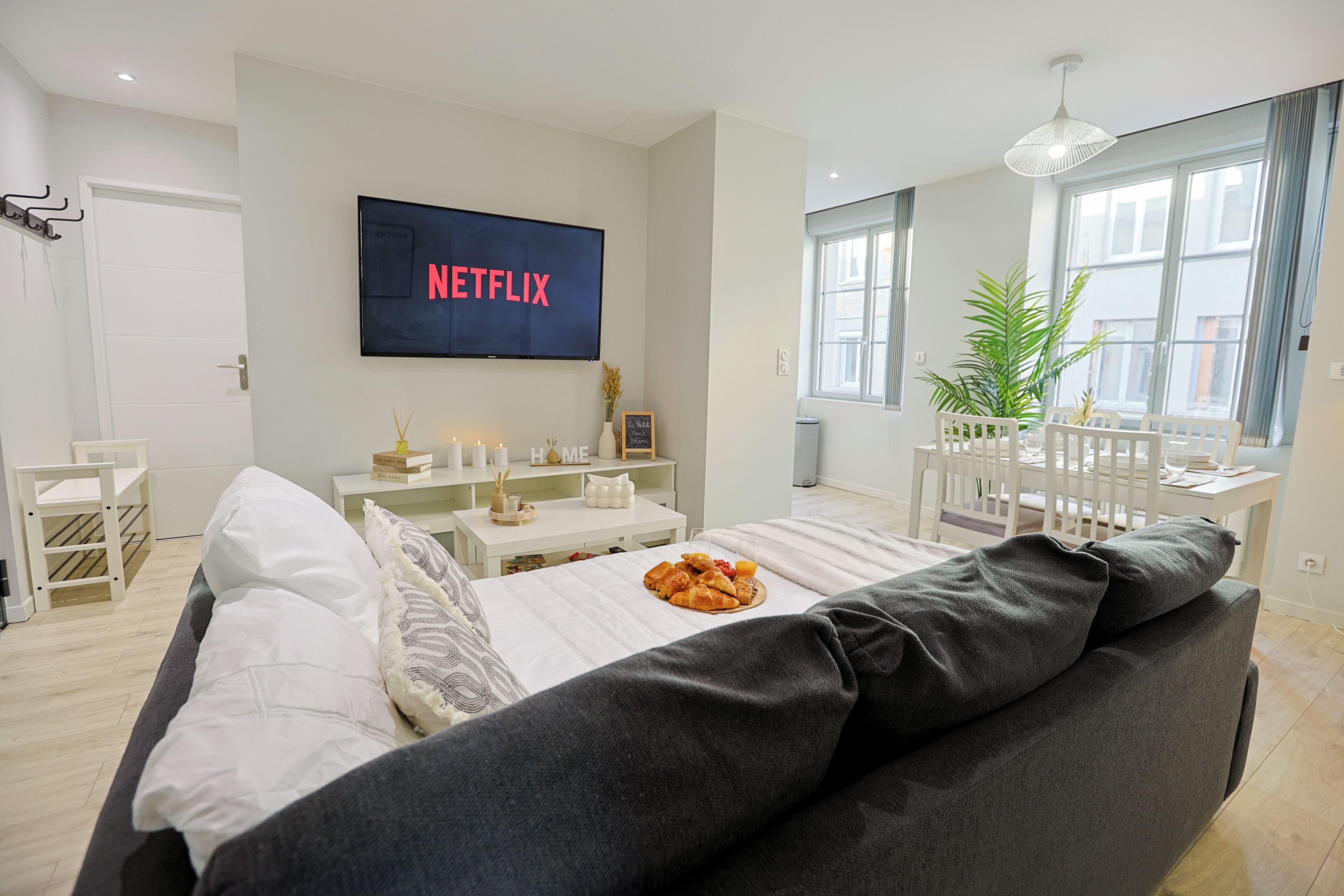 Una sala moderna con un televisor de pantalla plana grande mostrando 'NETFLIX', un sofá blanco, una mesa de comedor con sillas y una planta en maceta cerca de la ventana.