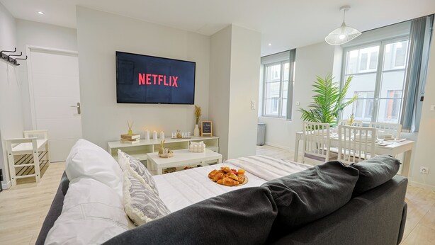 Un soggiorno moderno con un'ampia televisione a schermo piatto che mostra 'NETFLIX', un divano bianco, un tavolo da pranzo con sedie e una pianta in vaso vicino alla finestra.