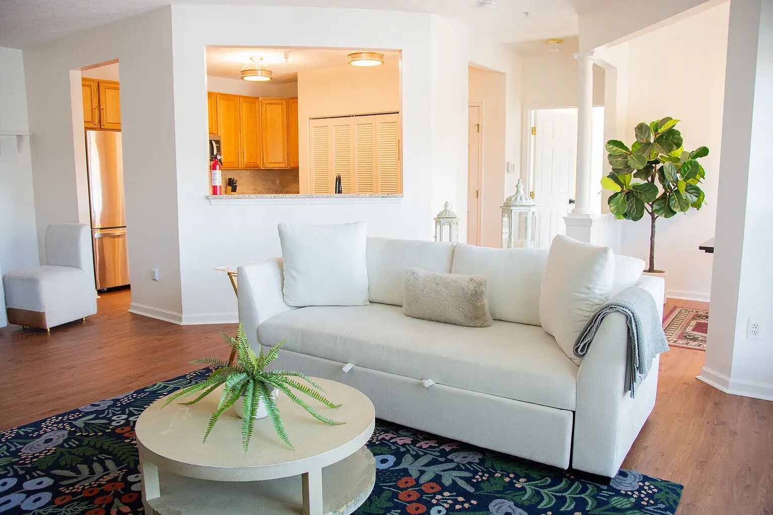 A living room with a sofa, a small round table with a plant, a patterned rug, and a view into the kitchen.
