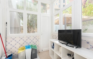 A room with a television on a white stand, a mop leaning against the wall, and a trash bin with colorful bags. - HOUZE Historical Lisbon, flat with private patio (Lisboa)