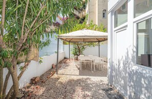 Ein überdachter Patio-Bereich mit Tisch und Stühlen, umgeben von einer weißen Mauer und einem Baum.