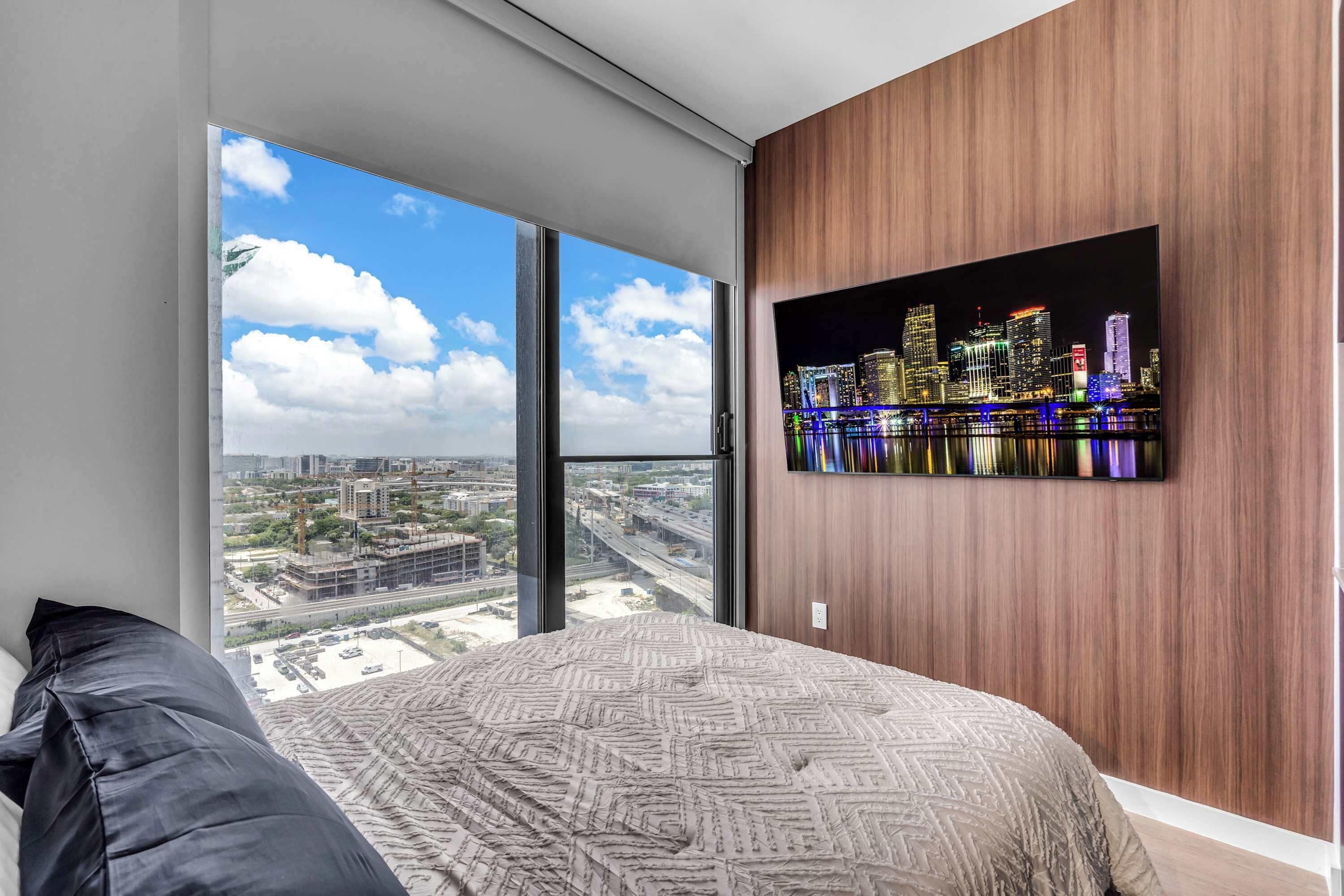Ein Schlafzimmer mit großem Fenster, einem Bett mit gemusterten Textilien und Blick auf die Stadt.