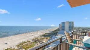 A beach view from a balcony with a clear sky, a sandy beach, and a high-rise building in the background. - Seaside Sanctuary - Beachfront (Orange Beach)