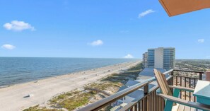 Blick auf einen Strand vom Balkon aus, mit blauem Himmel, Sandstrand und einem Hochhaus im Hintergrund.