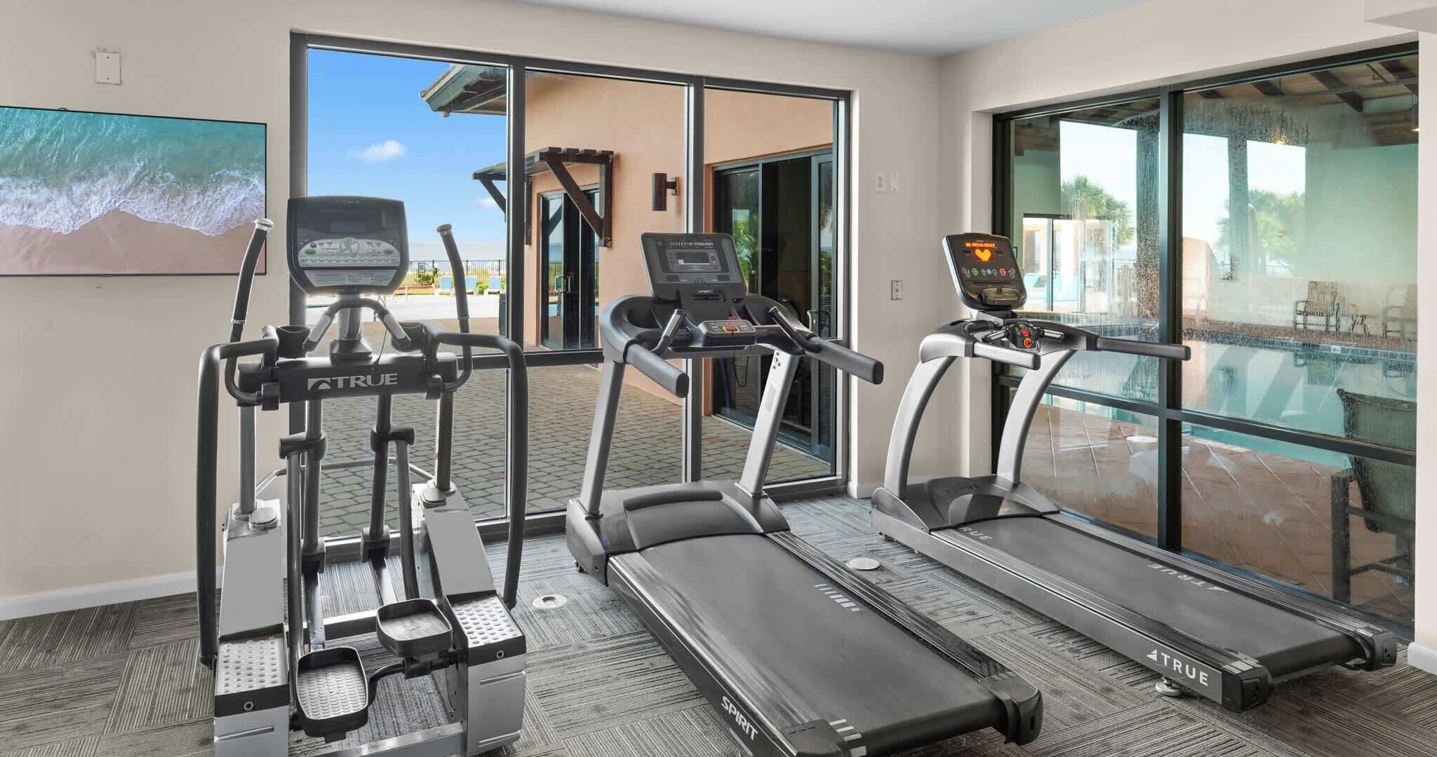 A fitness room with treadmills, a stair climber, and a stationary bike, with a pool visible through the glass doors.