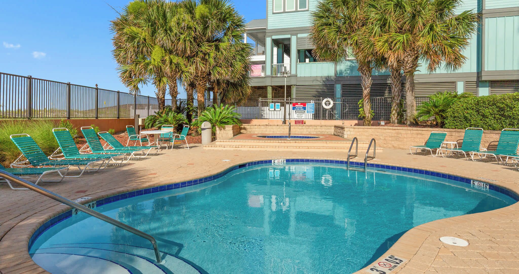 A swimming pool with lounge chairs, palm trees, and a building in the background.