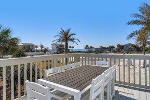 A wooden deck with white chairs and a table, overlooking a beach with palm trees and buildings in the background. - Sun and Sand (Panama City Beach)
