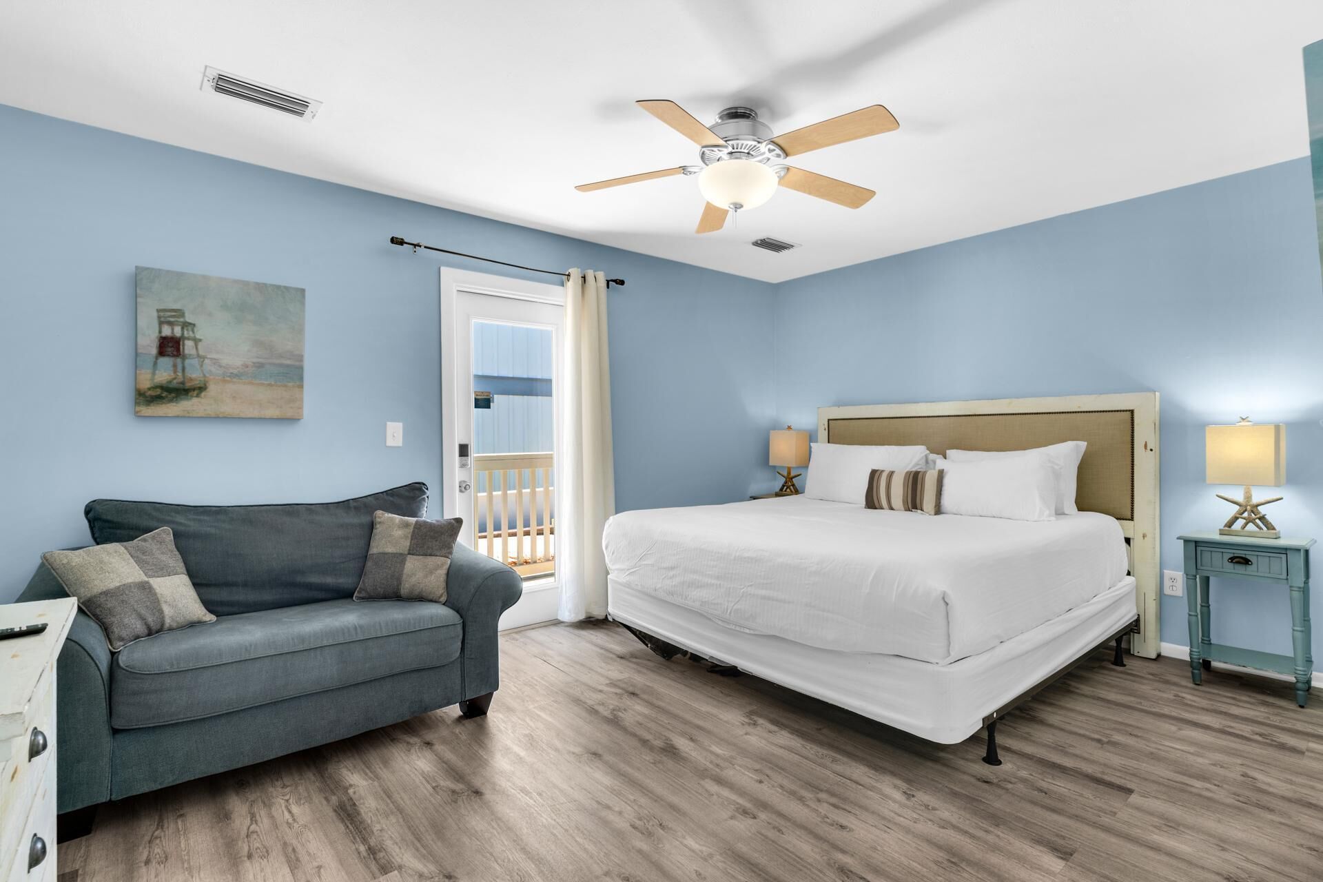 A hotel room with a bed, a sofa, a ceiling fan, and a painting on the wall.