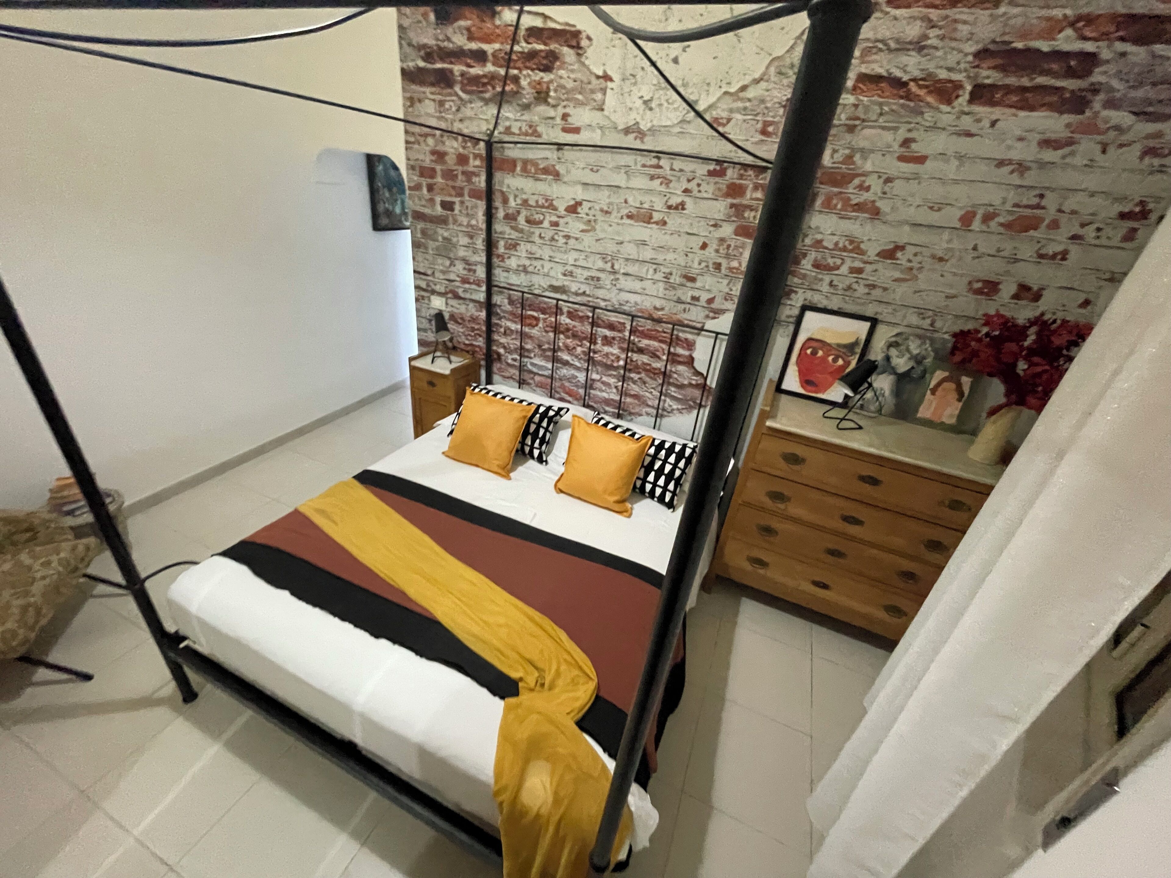 A four-poster bed with a striped bedspread, a nightstand with a lamp, and a brick wall backdrop.