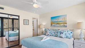 Ein Hotelzimmer mit einem Bett, einem Strandgemälde, einem Deckenventilator und Meerblick durch ein Fenster.