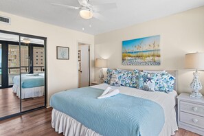 Ein Hotelzimmer mit einem Bett, einem Strandgemälde, einem Deckenventilator und Meerblick durch ein Fenster.