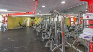 Ein Fitnessstudio mit verschiedenen Trainingsgeräten, einem Schild, das darauf hinweist, dass Gäste sich anmelden müssen, und einer Person, die im Hintergrund trainiert.