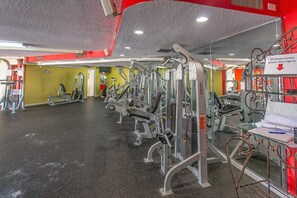 Ein Fitnessstudio mit verschiedenen Trainingsgeräten, einem Schild, das darauf hinweist, dass Gäste sich anmelden müssen, und einer Person, die im Hintergrund trainiert.