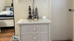 Ein Zimmer mit einem an der Wand befestigten Fernseher, einem Nachttisch aus Weidwerk mit Schubladen und einem Bett mit gestreifter Bettwäsche.