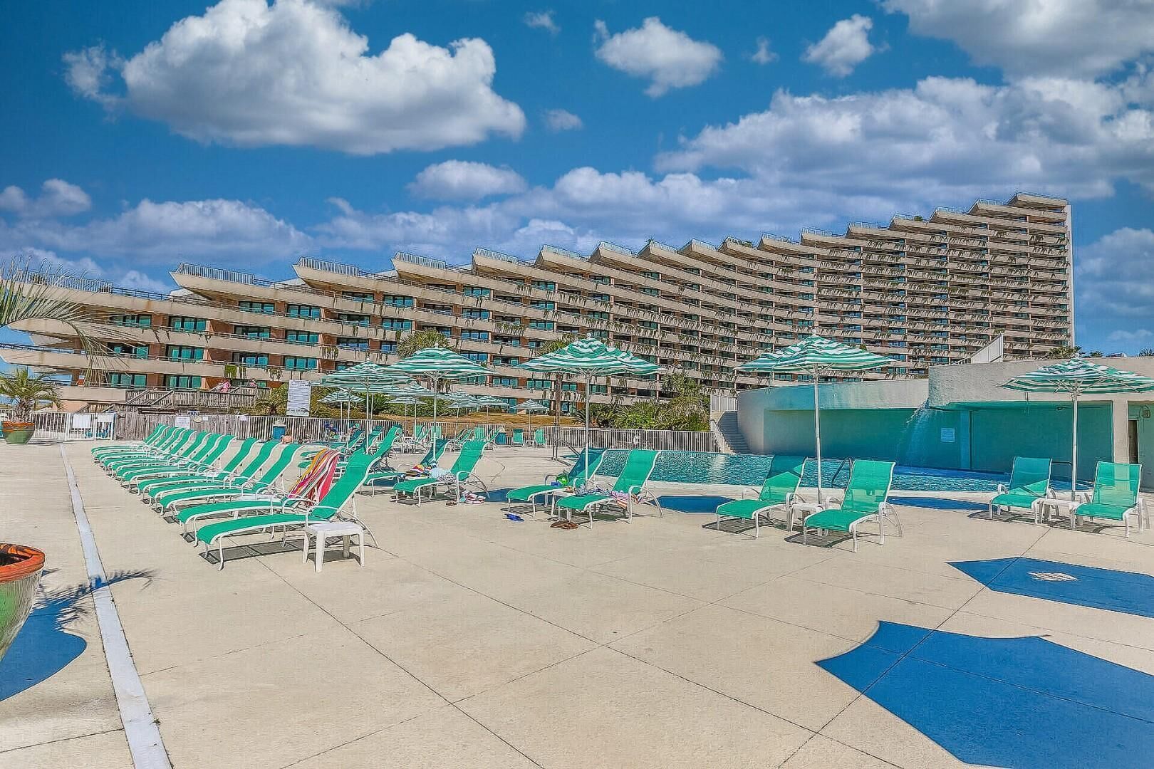 A hotel pool area with multiple lounge chairs and umbrellas, a large pool, and a high-rise building in the background.