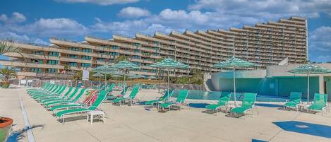 A hotel pool area with multiple lounge chairs and umbrellas, a large pool, and a high-rise building in the background.