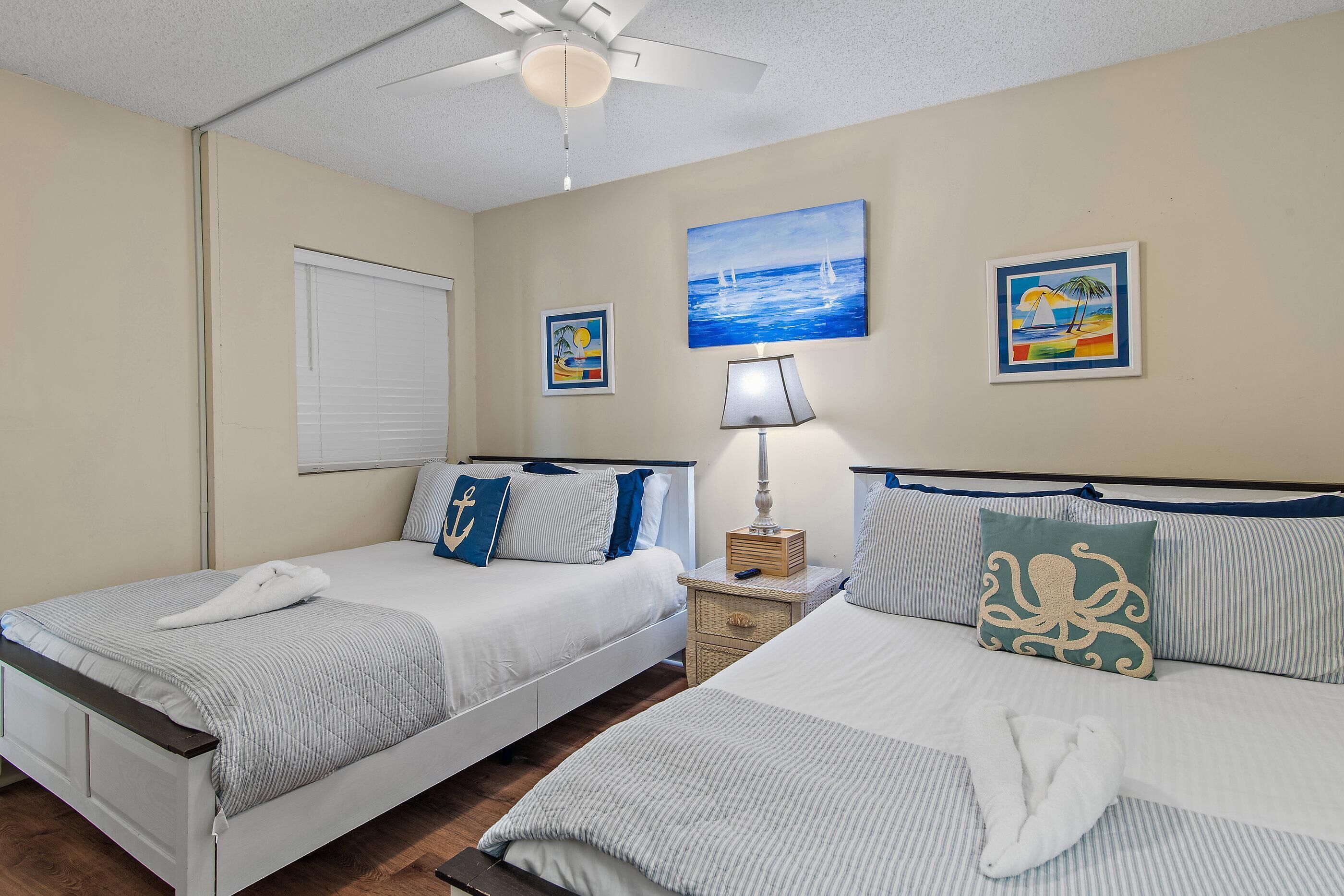 A hotel room with two beds, a ceiling fan, a painting, and a lamp.