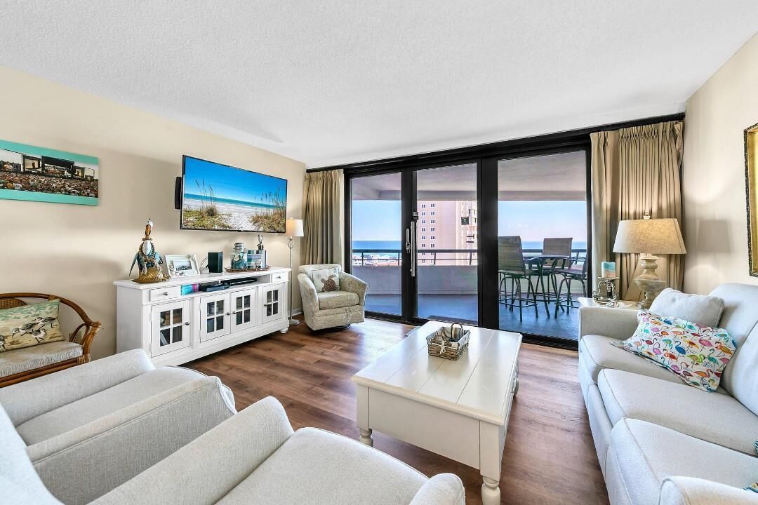 A hotel room with a balcony, a white sofa, a white armchair, a coffee table, and a TV on the wall.